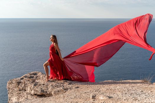 A girl with loose hair in a red dress stands on a rock rock above the sea. In the background, the sea and the rocks. The concept of travel.