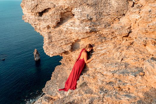 A girl with loose hair in a red dress stands on a rock rock above the sea. In the background, the sea and the rocks. The concept of travel.