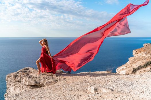 A girl with loose hair in a red dress stands on a rock rock above the sea. In the background, the sea and the rocks. The concept of travel.