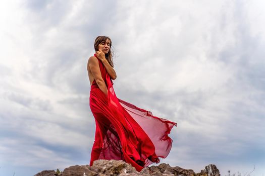 A woman in a red dress stands above a stormy sky, her dress fluttering, the fabric flying in the wind
