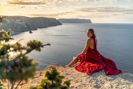 A girl with loose hair in a red dress sits on a rock rock above the sea. In the background, the sun rises from behind the mountains