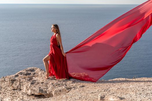 A girl with loose hair in a red dress stands on a rock rock above the sea. In the background, the sea and the rocks. The concept of travel.