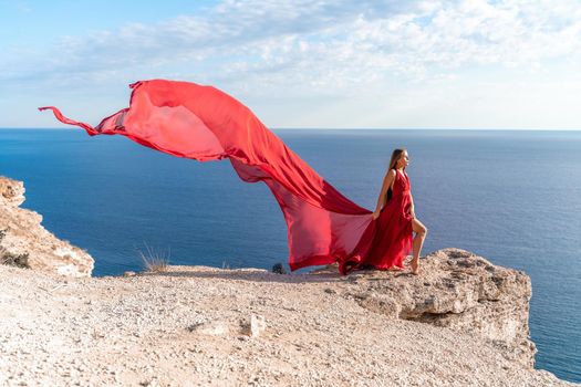 A girl with loose hair in a red dress stands on a rock rock above the sea. In the background, the sea and the rocks. The concept of travel.