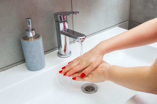 White sink with a silver faucet in the bathroom. Gray can with liquid soap for hands. Turning on the tap water, personal hand hygiene. Hand washing under running water