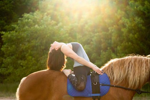 young woman doing yoga on a horse against the backdrop of trees.