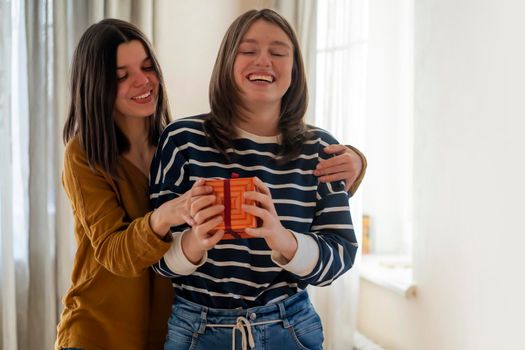 Two happy young girls laugh, friends hug and give each other gifts during the holiday. Women surprise and congratulate on the anniversary of their relationship in their cozy home.