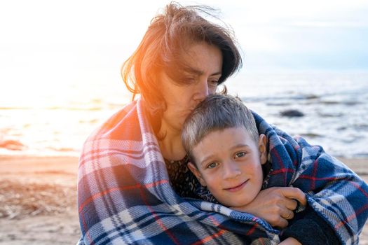 a happy mother and her preschooler son are relaxing on the beach wrapped in a plaid plaid from the wind in the light of the setting sun. parental love. family ties. Mother's Day
