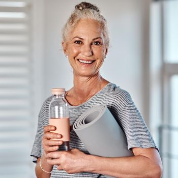 I have everything I need to get started with my workout. Portrait of a cheerful mature woman holding a bottle of water and yoga mat ready to start her morning session of yoga inside of a studio