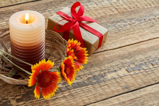 Burning candle, gift box wrapped in kraft paper with red ribbon and flowers on wooden boards. Top view. Holiday concept.