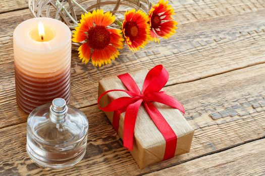 Perfume, gift or present box wrapped in kraft paper with red ribbon, burning candle and yellow flowers on wooden boards. Top view. Holiday concept.