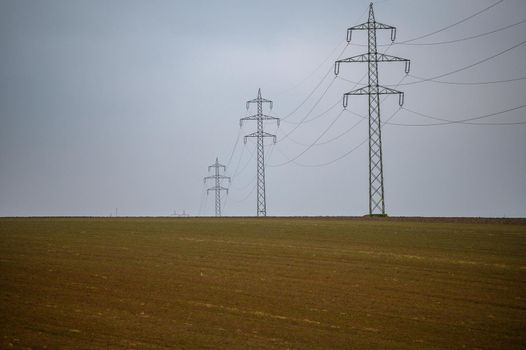 A high-voltage line runs over a bald field. The gray clouds of fog reinforce the wintry impression of the landscape.