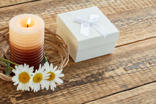 Burning candle, bouquet of chamomile flowers and white gift or present box on wooden boards. Top view. Holiday concept.