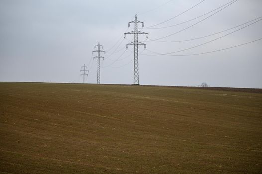 A high-voltage line runs over a bald field. The gray clouds of fog reinforce the wintry impression of the landscape.