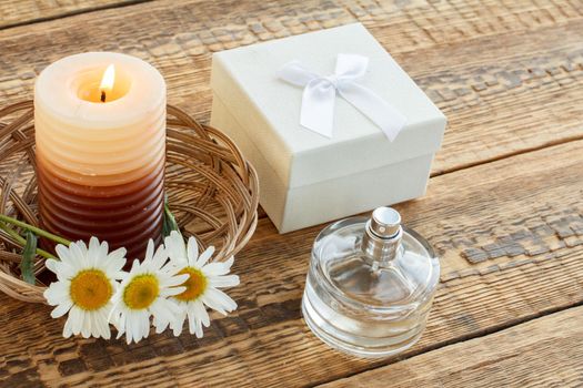 Bouquet of chamomiles, perfume, burning candle and white gift or present box on wooden boards. Top view. Holiday concept.