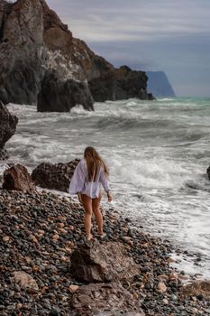 A beautiful girl in a white shirt and black swimsuit stands on the edge of a cliff, big waves with white foam. A cloudy stormy day at sea, with clouds and big waves hitting the rocks