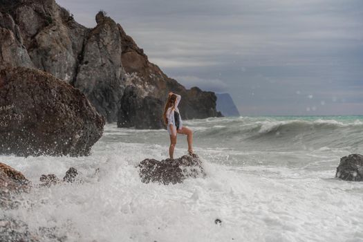 A beautiful girl in a white shirt and black swimsuit stands on a rock, big waves with white foam. A cloudy stormy day at sea, with clouds and big waves hitting the rocks