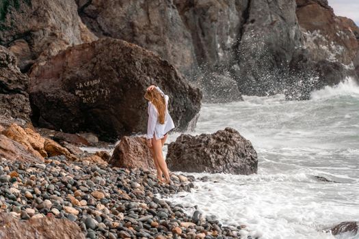 A beautiful girl in a white shirt and black swimsuit stands on the edge of a cliff, big waves with white foam. A cloudy stormy day at sea, with clouds and big waves hitting the rocks