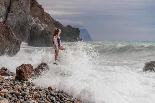 A beautiful girl in a white shirt and black swimsuit stands on a rock, big waves with white foam. A cloudy stormy day at sea, with clouds and big waves hitting the rocks
