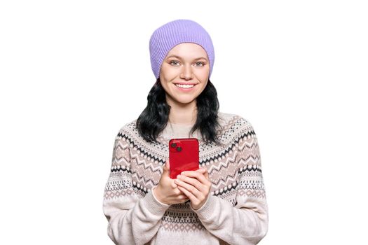 Beautiful young teenage girl in hat in a woolen sweater with smartphone in her hands looking at camera on white isolated background. Winter season, Christmas and New Year holidays concept