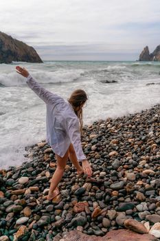 A beautiful girl in a black dress is walking on the waves, big waves with white foam. A cloudy stormy day at sea, with clouds and big waves hitting the rocks