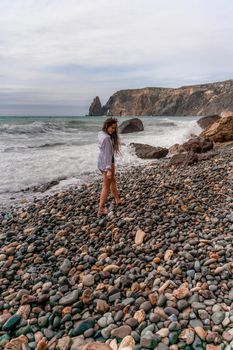 A beautiful girl in a black dress is walking on the waves, big waves with white foam. A cloudy stormy day at sea, with clouds and big waves hitting the rocks