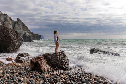 A beautiful girl in a white shirt and black swimsuit stands on a rock, big waves with white foam. A cloudy stormy day at sea, with clouds and big waves hitting the rocks