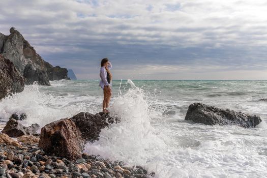 A beautiful girl in a white shirt and black swimsuit stands on a rock, big waves with white foam. A cloudy stormy day at sea, with clouds and big waves hitting the rocks