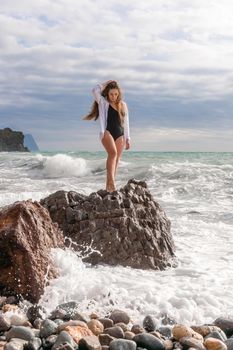 A beautiful girl in a white shirt and black swimsuit stands on a rock, big waves with white foam. A cloudy stormy day at sea, with clouds and big waves hitting the rocks