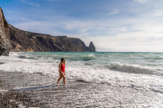 A beautiful and sexy brunette in a red swimsuit on a pebble beach, Running along the shore in the foam of the waves.