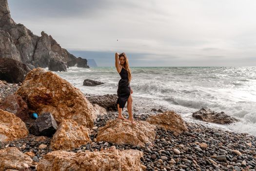 A beautiful girl in a black dress stands on a rock, big waves with white foam. A cloudy stormy day at sea, with clouds and big waves hitting the rocks