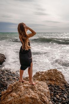 A beautiful girl in a black dress stands on a rock, big waves with white foam. A cloudy stormy day at sea, with clouds and big waves hitting the rocks