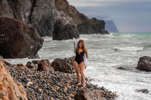 A beautiful girl in a white shirt and black swimsuit stands on the edge of a cliff, big waves with white foam. A cloudy stormy day at sea, with clouds and big waves hitting the rocks