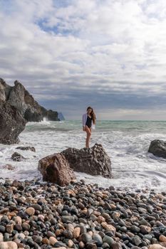 A beautiful girl in a white shirt and black swimsuit stands on a rock, big waves with white foam. A cloudy stormy day at sea, with clouds and big waves hitting the rocks