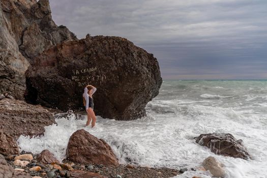 A beautiful girl in a black dress is walking on the waves, big waves with white foam. A cloudy stormy day at sea, with clouds and big waves hitting the rocks