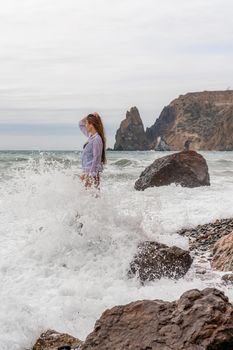 A beautiful girl in a black dress is walking on the waves, big waves with white foam. A cloudy stormy day at sea, with clouds and big waves hitting the rocks