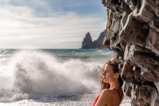 A beauty in a red swimsuit with long legs poses on a fantastic beach with huge waves against the background of mountains.