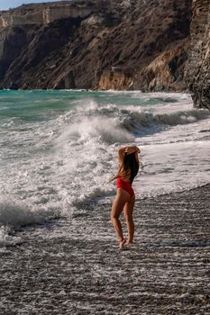 A beautiful and sexy brunette in a red swimsuit on a pebble beach, Running along the shore in the foam of the waves.