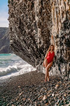 A beauty in a red swimsuit with long legs poses on a fantastic beach with huge waves against the background of mountains.