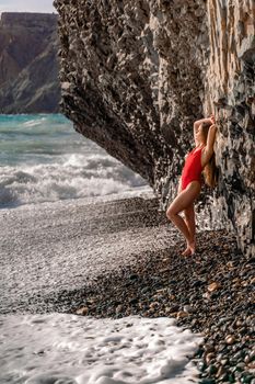 A beauty in a red swimsuit with long legs poses on a fantastic beach with huge waves against the background of mountains.