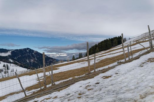 A snow-covered steep alpine meadow with a wooden fence. The deep blue sky is partially covered and announces the next snowstorm.