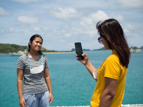 A woman in a yellow shirt is holding a phone to take pictures of a girl in a gray shirt. With a background of blue water and small mountains. It is tourism after the corona virus outbreak.