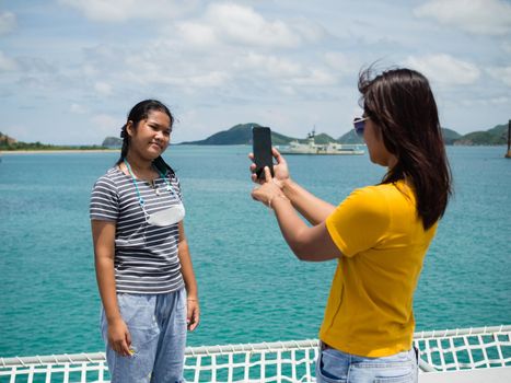 A woman in a yellow shirt is holding a phone to take pictures of a girl in a gray shirt. With a background of blue water and small mountains. It is tourism after the corona virus outbreak.