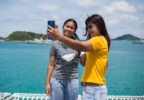 A woman in a yellow shirt is holding a phone to take a selfie with a girl in a gray shirt. with a background of blue water and small mountains. It is tourism after the corona virus outbreak.