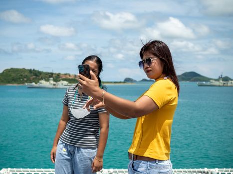 A woman in a yellow shirt is holding a phone to take a selfie with a girl in a gray shirt. with a background of blue water and small mountains. It is tourism after the corona virus outbreak.
