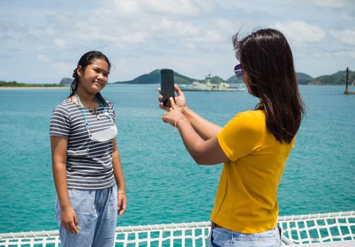 A woman in a yellow shirt is holding a phone to take pictures of a girl in a gray shirt. With a background of blue water and small mountains. It is tourism after the corona virus outbreak.