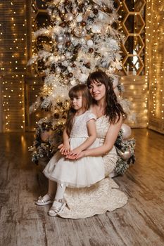 A little girl with her mother in light festive dresses next to the Christmas tree. The theme of New Year's holidays and festive interior with garlands and light bulbs.
