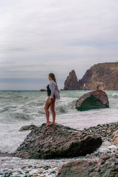 A beautiful girl in a white shirt and black swimsuit stands on a rock, big waves with white foam. A cloudy stormy day at sea, with clouds and big waves hitting the rocks