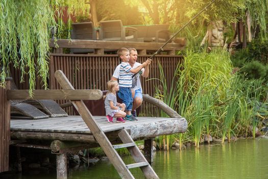 The distant plan, dad teaches his son to fish, the girl is watching them. Sunny warm summer day. Beautiful greenery around a small lake. The concept of a happy. International Day of Families