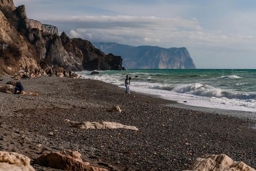 A beautiful girl in a white shirt and black swimsuit stands on the edge of a cliff, big waves with white foam. A cloudy stormy day at sea, with clouds and big waves hitting the rocks