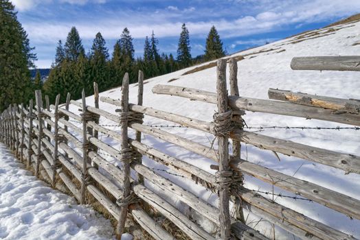 An old wooden fence that is held together with willow rods and separates two snowy pastures from each other. In addition, the fence is reinforced with a barbed wire.